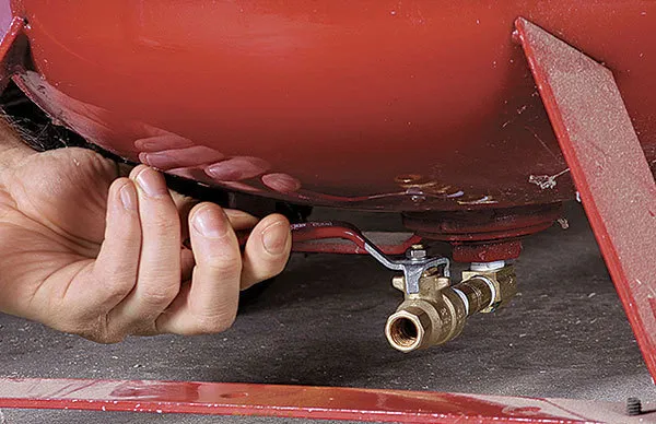 Compressor de ar sendo drenado manualmente pela válvula inferior para remover água acumulada e evitar corrosão interna conforme normas de segurança.
