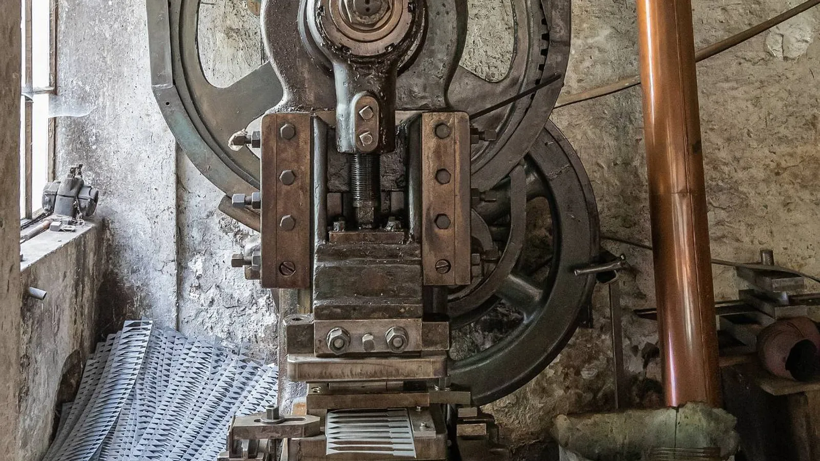 Prensa excêntrica antiga com volante, engrenagem e martelo expostos, pedal no piso e retalhos de chapa ao redor, preservada em oficina histórica