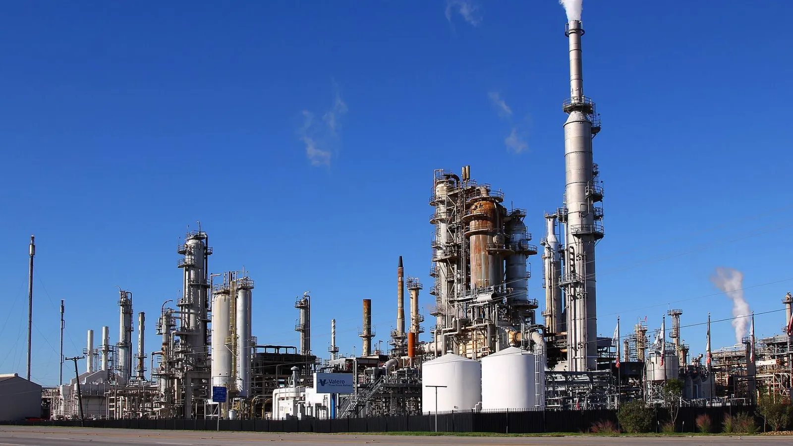 Refinaria com torres, vasos de pressão, tanques e chaminés sob céu azul, exemplo de planta com grande parque de equipamentos