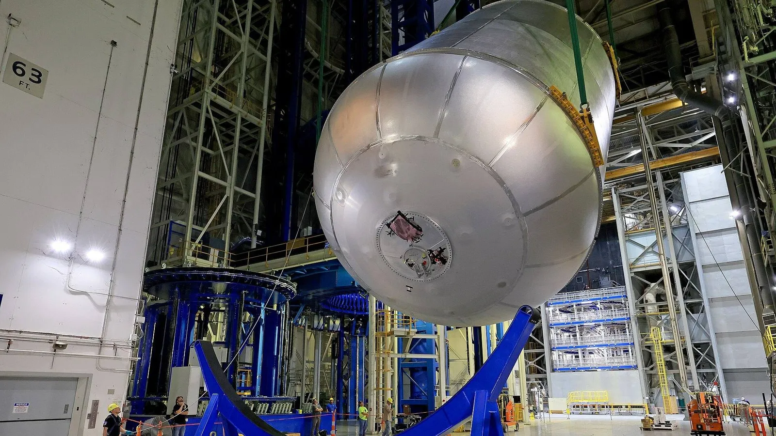 Grande tanque metálico suspenso por cabos sobre berço de transporte azul em galpão industrial, com equipe acompanhando a movimentação após o teste de prova