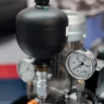 Pequeno vaso de pressão preto com dois manômetros de mostrador em unidade hidráulica exposta em feira industrial